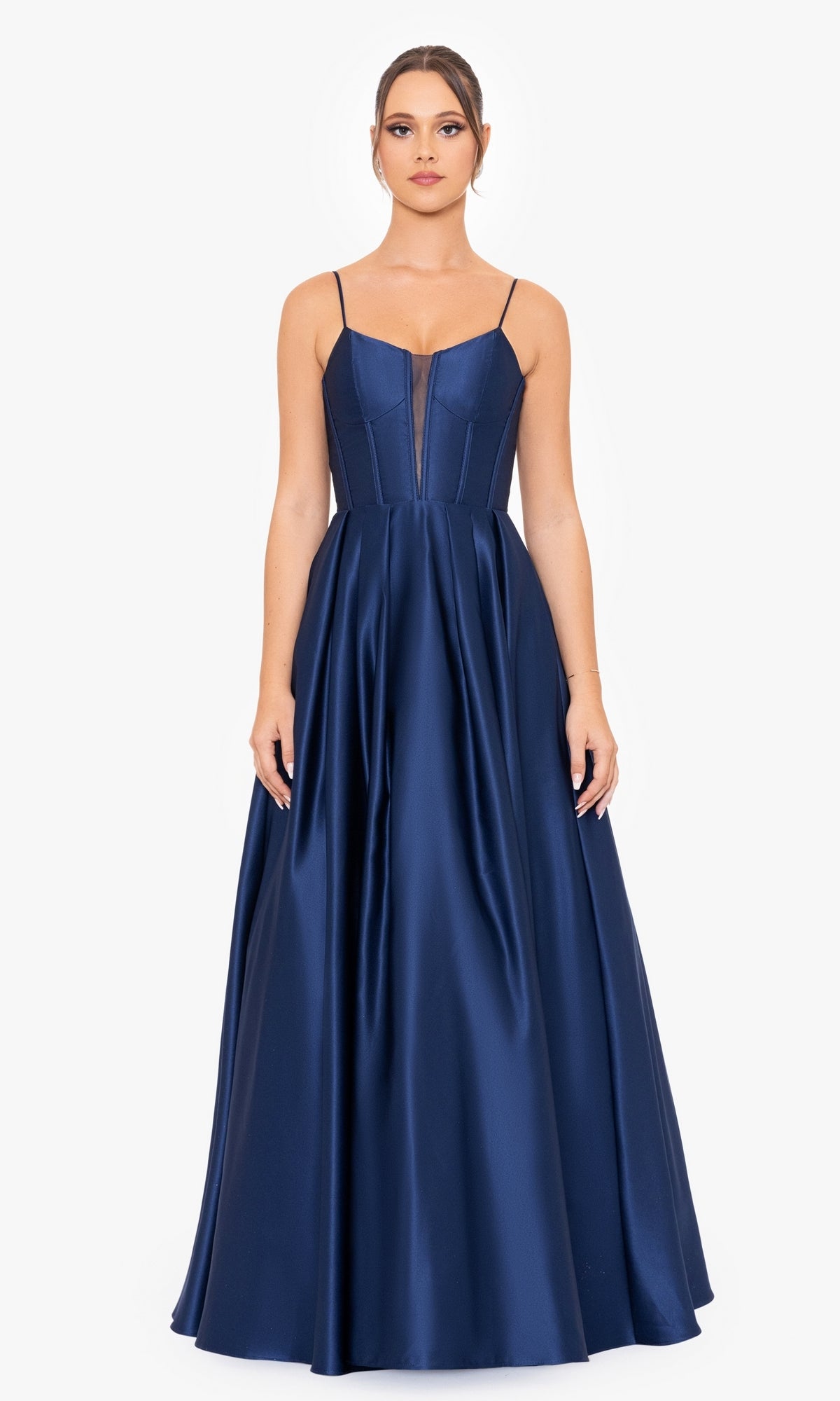 A woman models the Blondie Nites Long A-Line Prom Dress 4826BN—a sleeveless, floor-length navy satin gown with a corset-style bodice and spaghetti straps—standing against a plain white background.